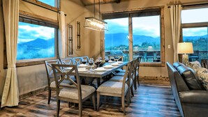 Un coin repas avec une longue table dressée pour six personnes, de grandes fenêtres offrant une vue sur les montagnes et un canapé en cuir dans un coin.
