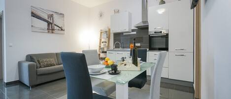 A modern kitchen with white cabinets, a dining table with chairs, a sofa, and a pendant light.
