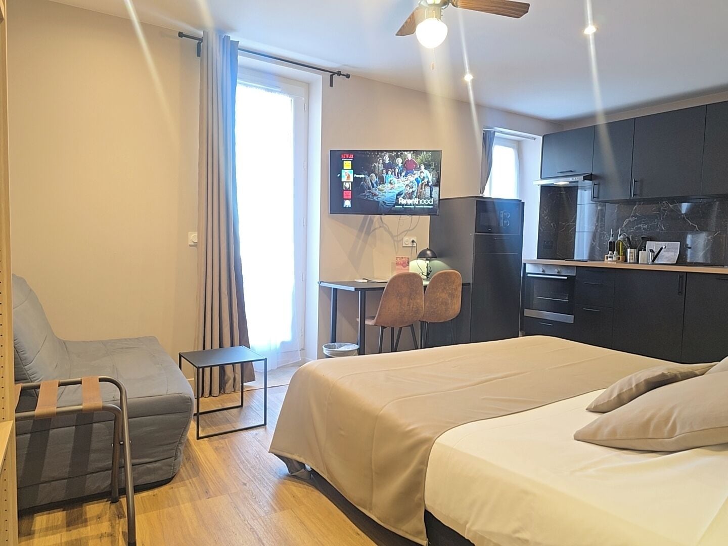 A modern hotel room with a bed, a desk, a chair, a TV, and a kitchenette.