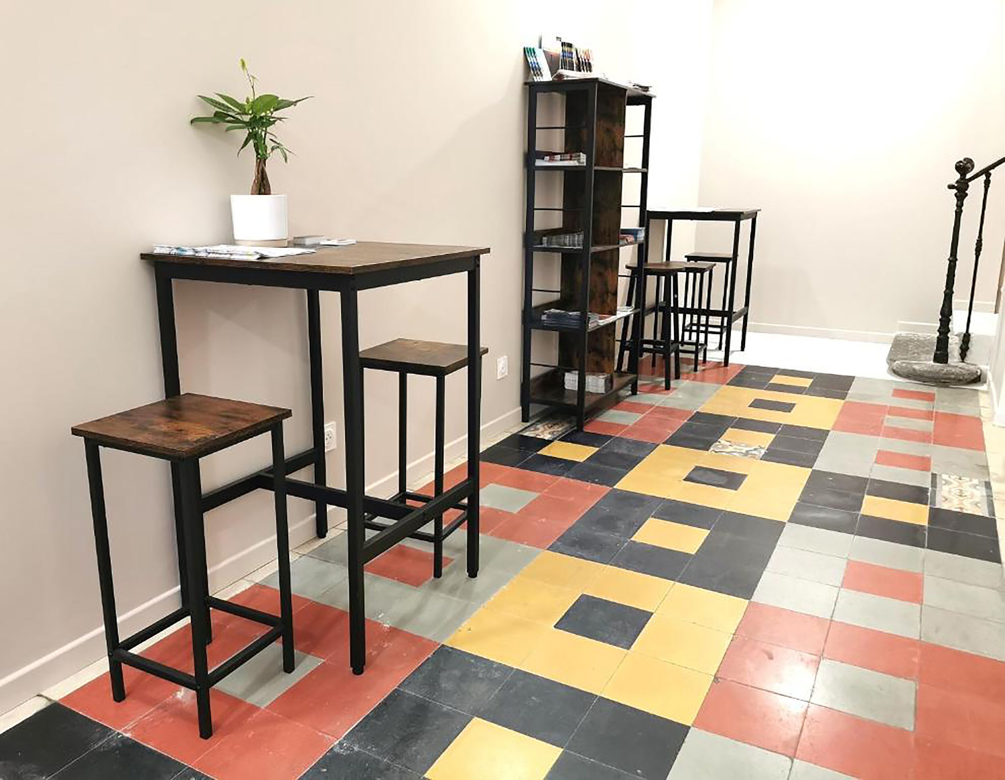 A hallway with a checkered floor, a black metal table with stools, a bookshelf, and a potted plant.