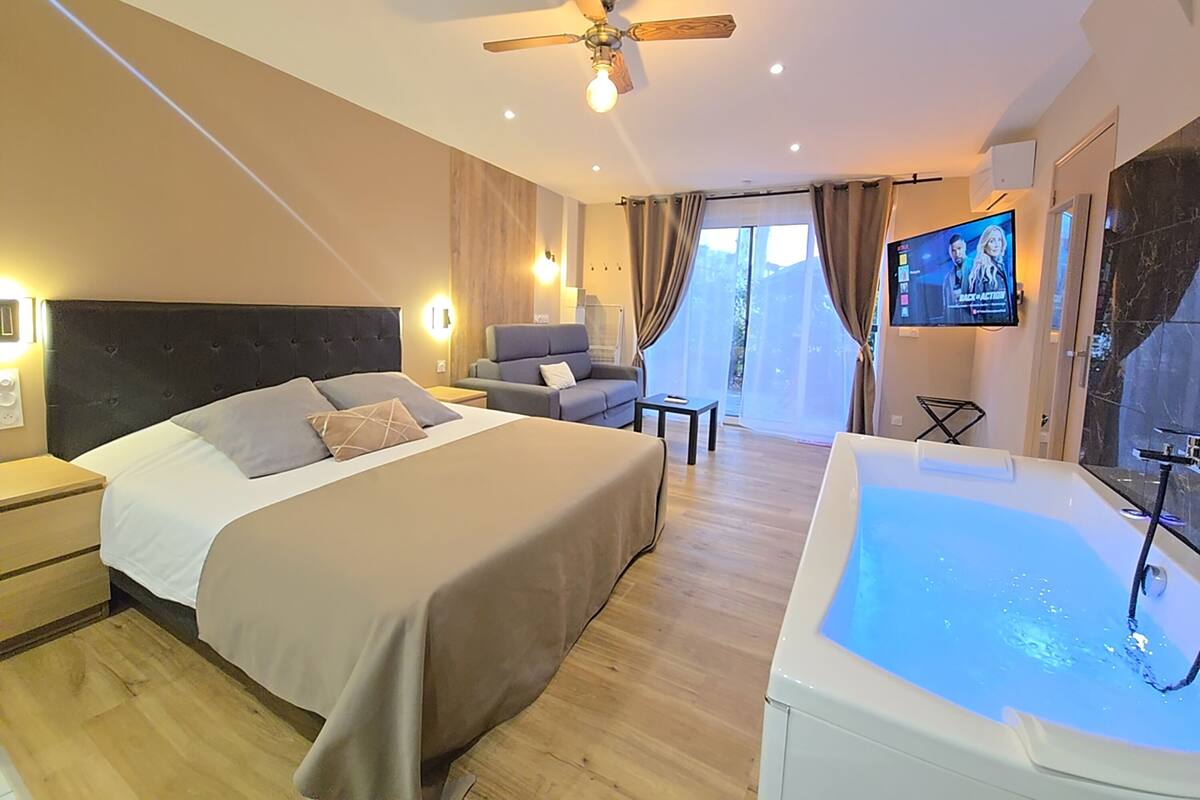A modern hotel room with a large bed, a sitting area, a TV, and a bathtub.