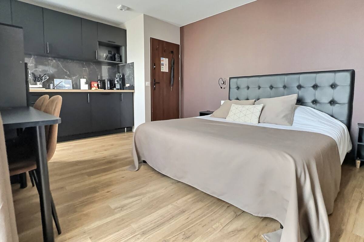 A modern bedroom with a large bed, a desk, and a kitchenette.