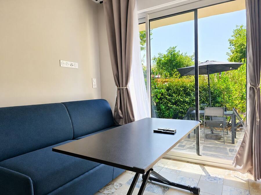 A room with a blue sofa, a small table, a window with curtains, and a view of greenery outside.