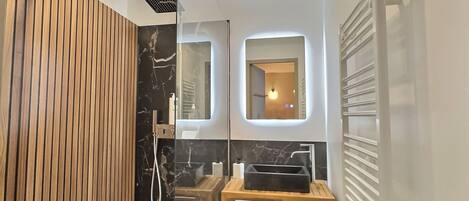 A modern bathroom with a wooden panel wall, a glass shower enclosure, a marble backsplash, a sink with a mirror, and a towel rack.