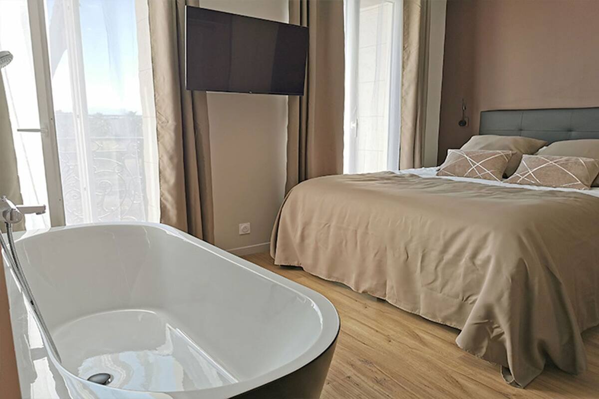 A bedroom with a large bathtub, a bed with beige bedding, and a television mounted on the wall.