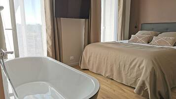 Une chambre avec une grande baignoire, un lit avec une literie beige et un téléviseur fixé au mur.