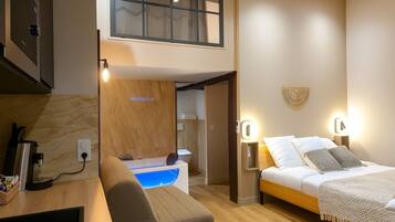 A modern hotel room with a bed, bedside table, and a built-in heater. There is a small seating area with a bench and a table, and a window with a view of the outside.