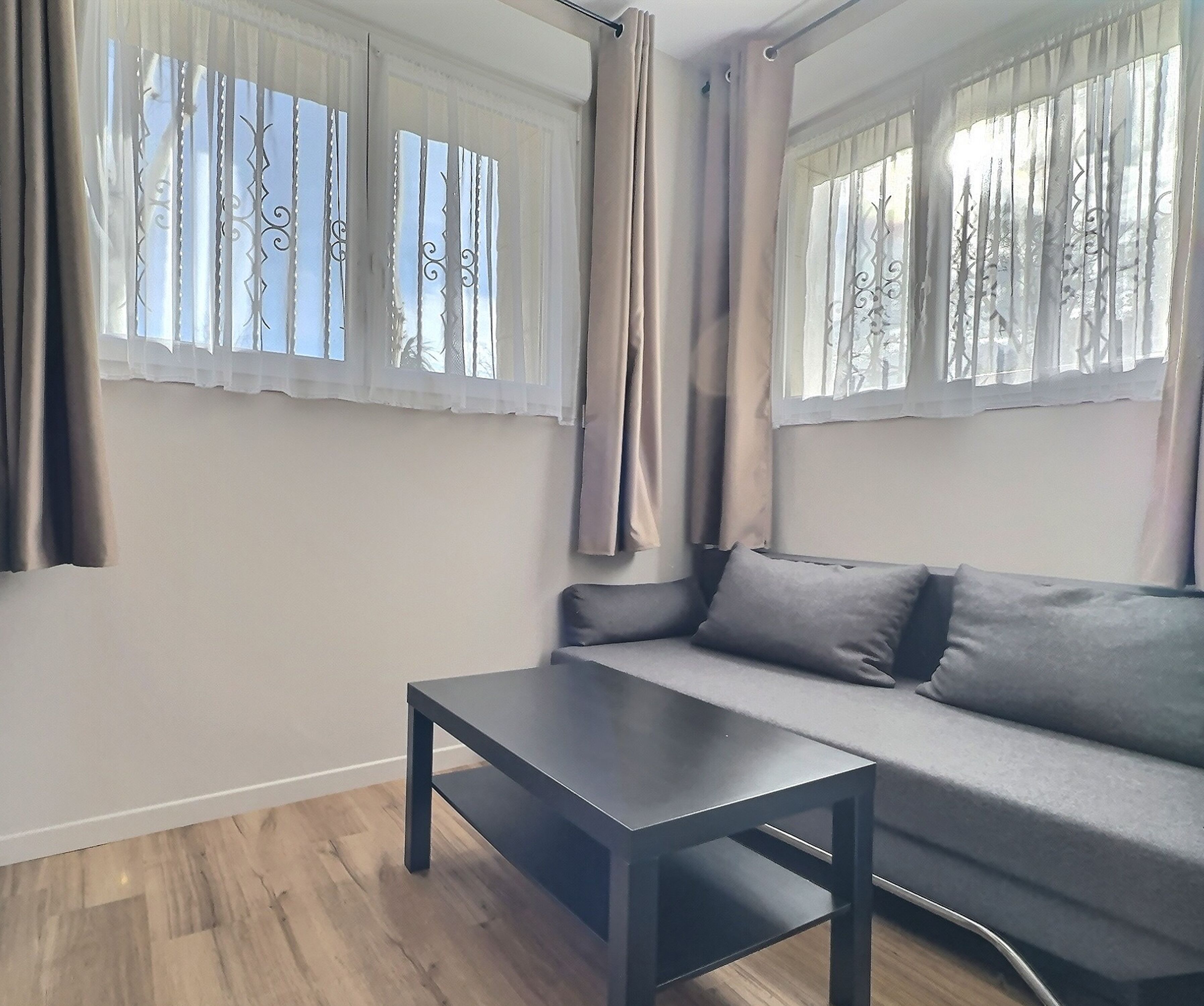 A modern living room with a grey sofa, a dark wooden coffee table, and a window with decorative curtains.