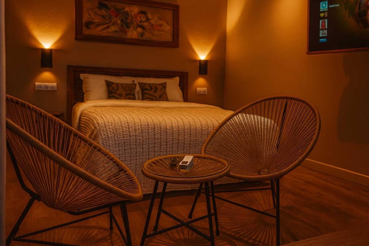 A hotel room with a bed, two wicker chairs, a small table, and wall-mounted lamps.