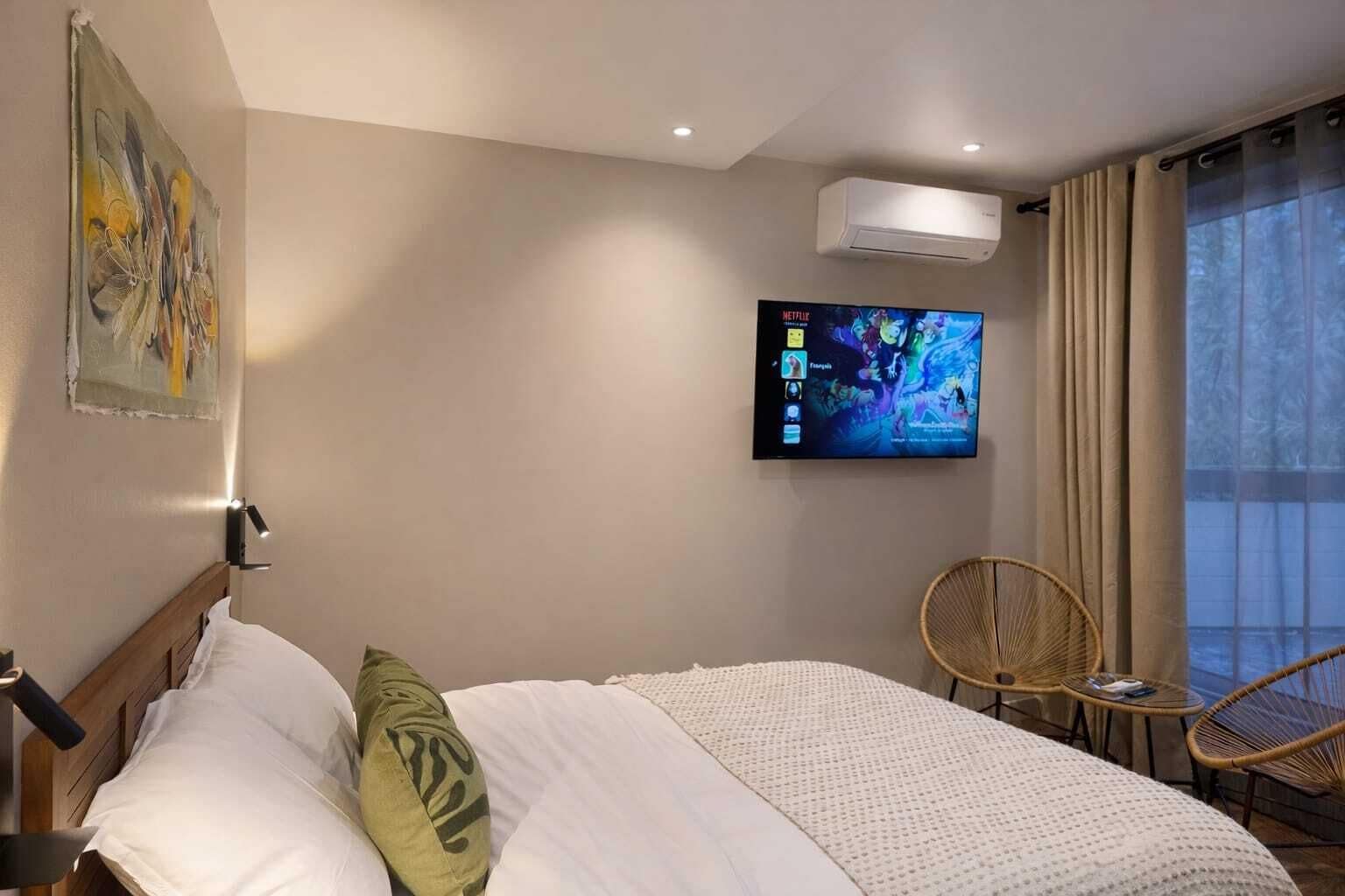 A hotel room with a bed, a TV mounted on the wall, a chair, and a window with curtains.