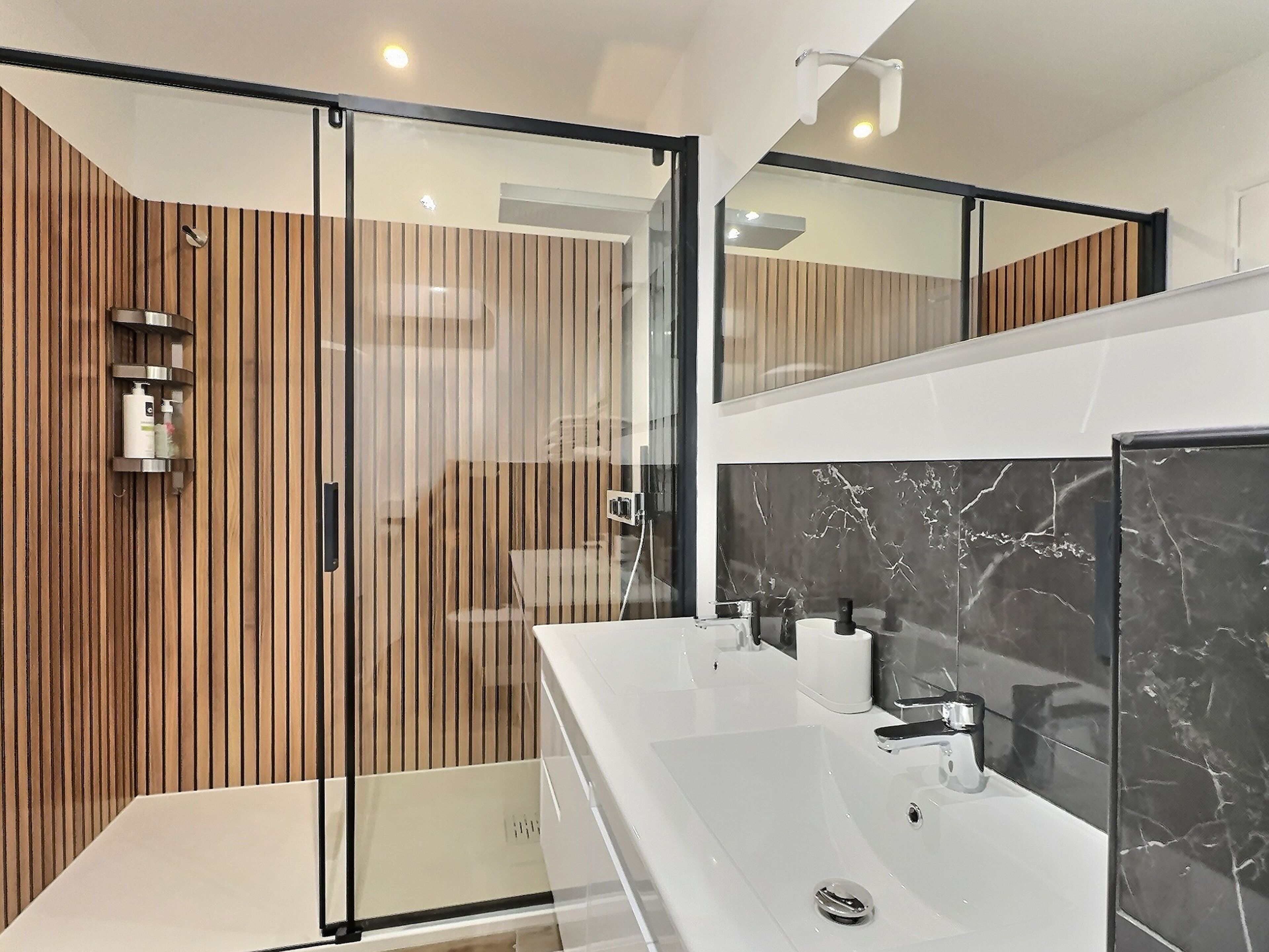 Un baño moderno con cabina de ducha de vidrio, paneles de madera, un tocador blanco y una salpicadura de mármol, y un espejo grande.