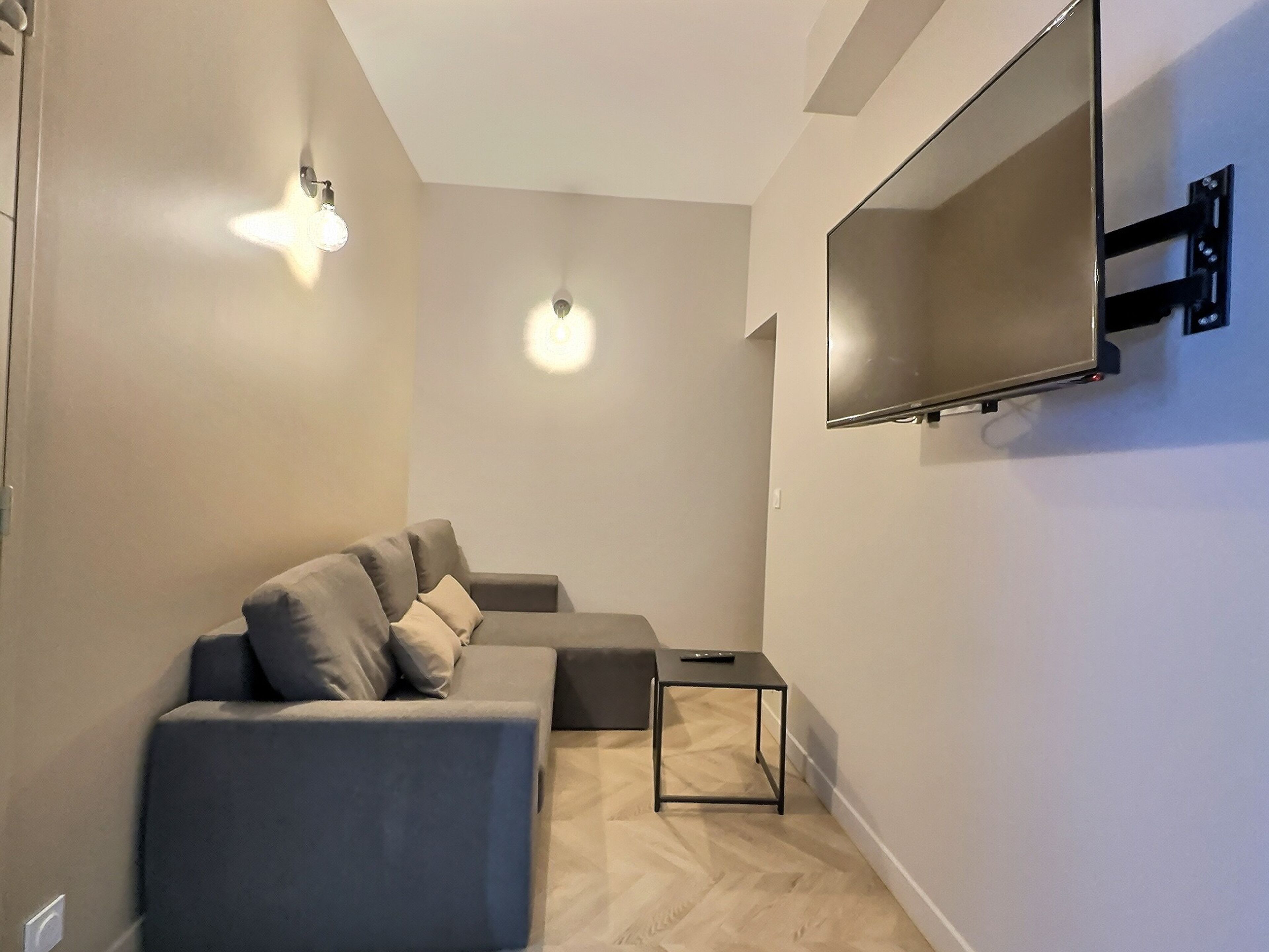 Un salon moderne avec un canapé d’angle gris, une petite table d’appoint noire et un téléviseur fixé au mur.