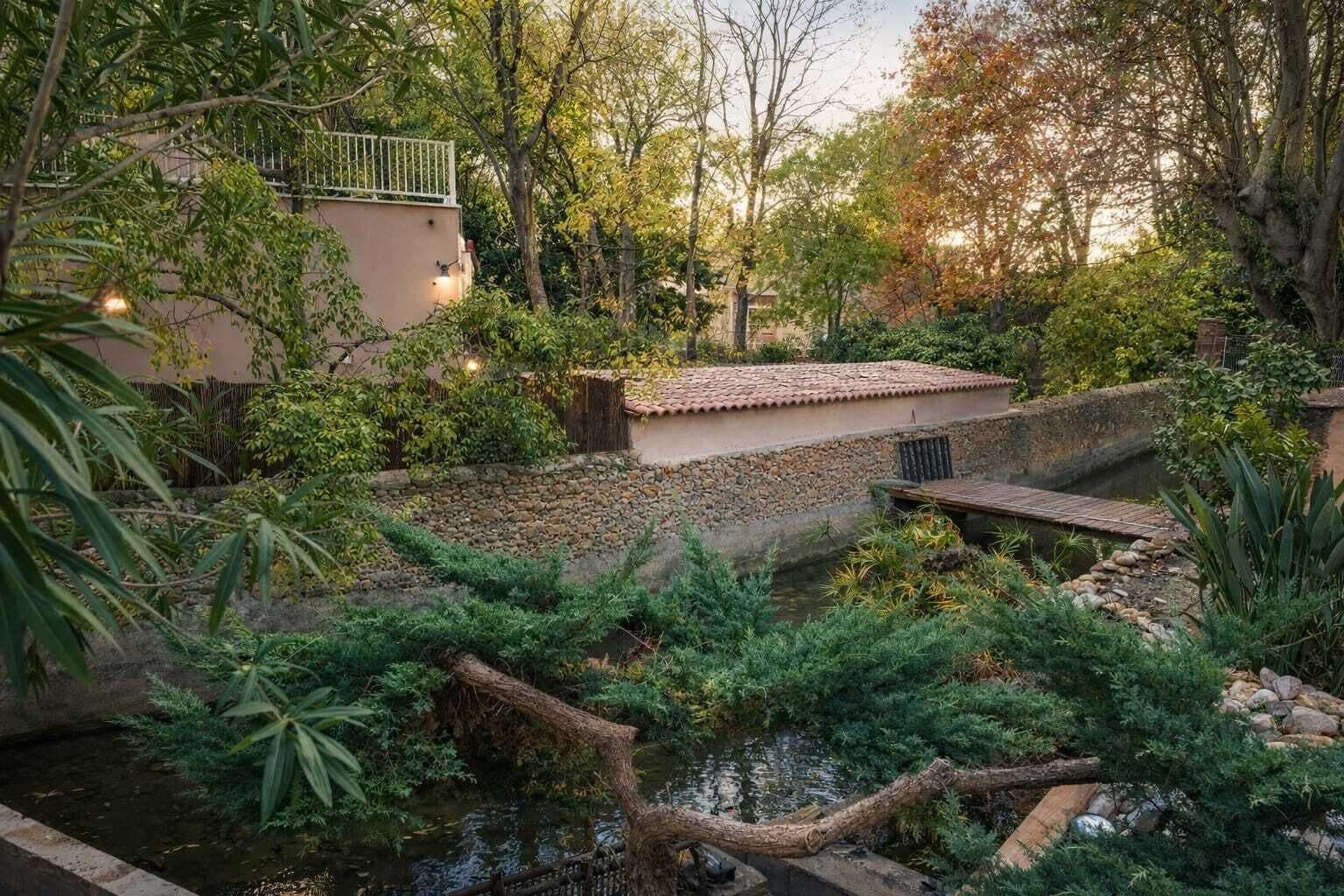 A serene garden with a small stream, a stone bridge, and a building with a tiled roof.