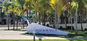 Una gran escultura blanca con el texto "PLAYA BLANCA" en un espacio público al aire libre, con palmeras y un sendero pavimentado.