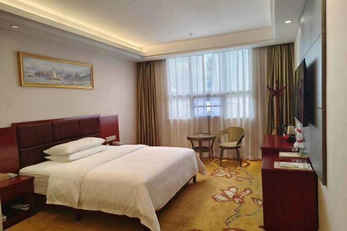 A hotel room with a large bed, a desk, a chair, a TV, and a window with curtains.