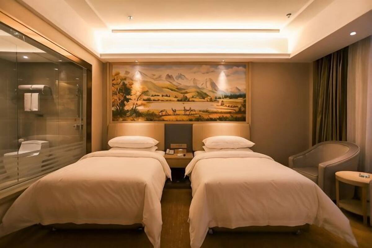 A hotel room with two beds, a painting of a landscape, a chair, and a glass shower enclosure.