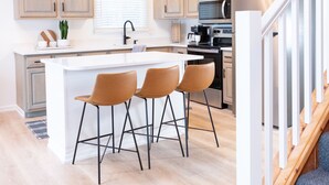 Una cocina moderna con armarios de madera, una isla central con taburetes de bar, electrodomésticos de acero inoxidable y una escalera con barandilla de madera.