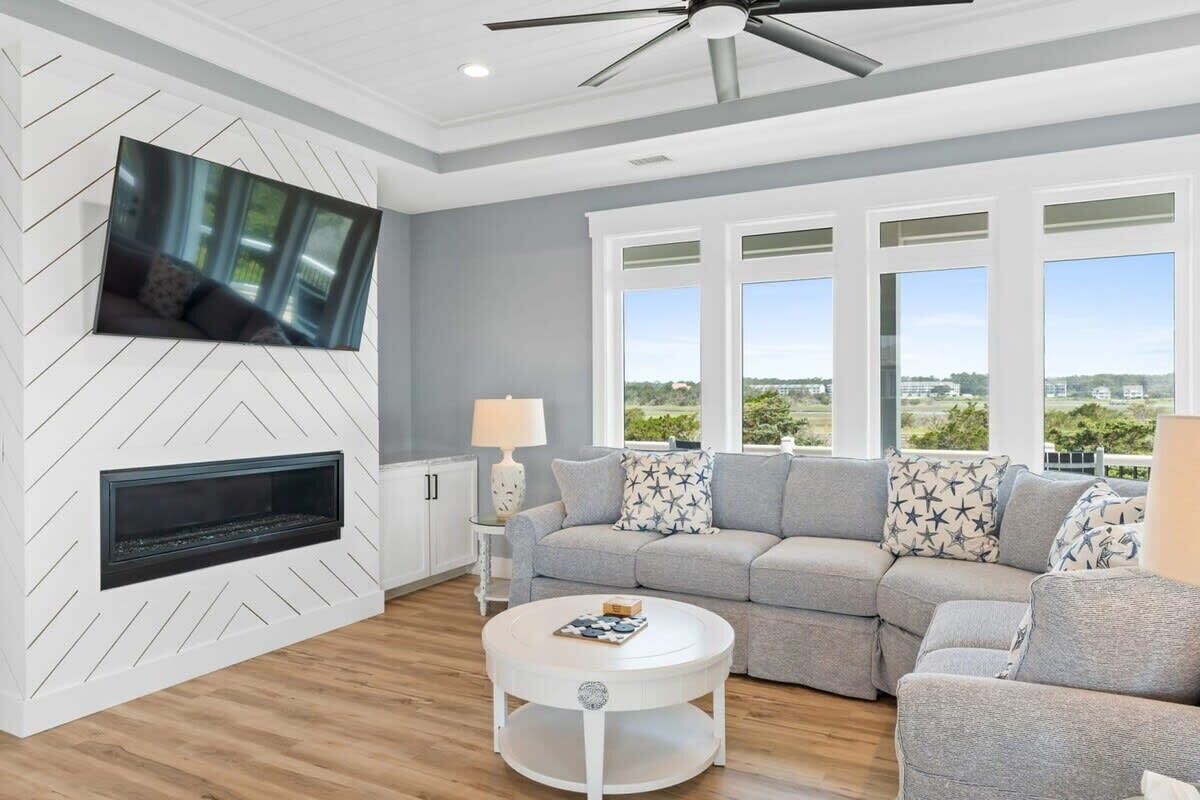 A modern living room with a fireplace, grey sofa set, white coffee table, and a large TV mounted on the wall.