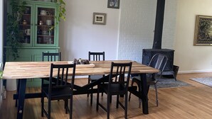 Ruokailutilassa on puinen pöytä ja tuolit, vihreä kaappi lasiovilla sekä musta takka.