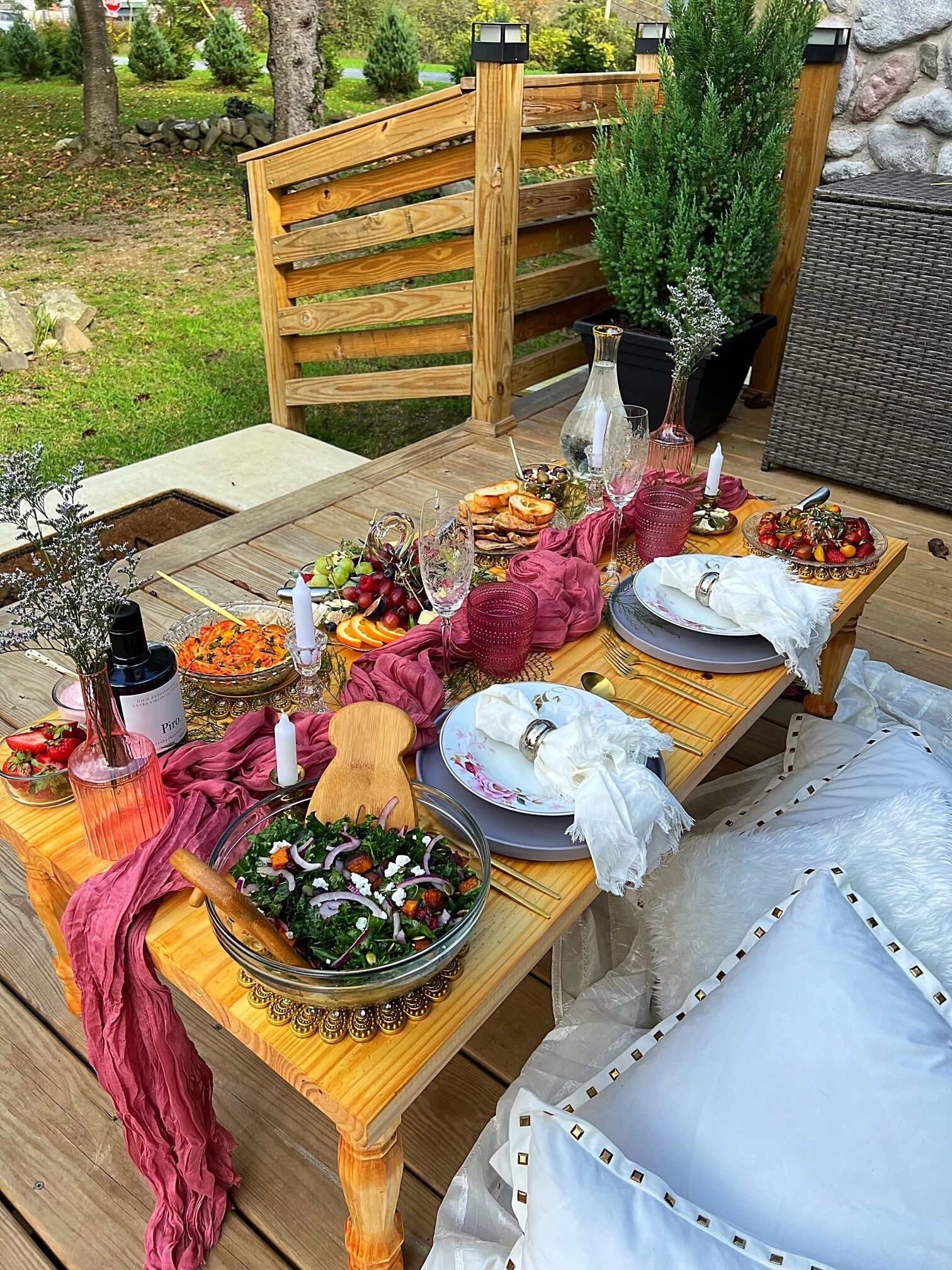 Een buitentafel gedekt met verschillende gerechten, glazen en een fles wijn, omringd door een houten hek en groen.