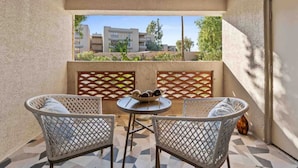 A balcony with two wicker chairs, a small round table with a bowl of fruits, and a view of buildings and greenery.