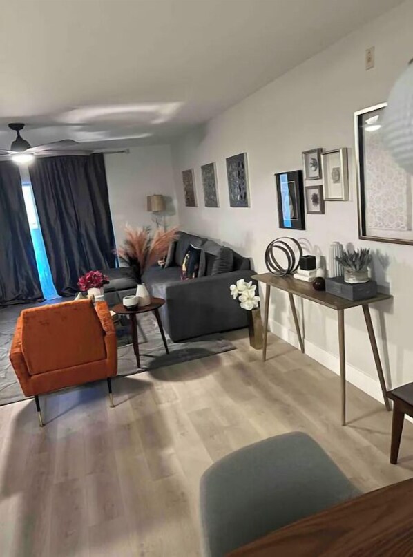 A modern living room with a grey sofa, a small orange chair, and a blue armchair. There is a coffee table with a vase of flowers and a lamp. The room has a wooden floor and a ceiling fan.