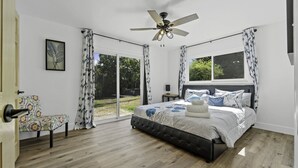 Un dormitorio con cama, una silla, ventilador de techo y ventanas amplias.