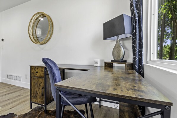 A room with a wooden desk, a chair, a lamp, a mirror, and a view of trees through a window.