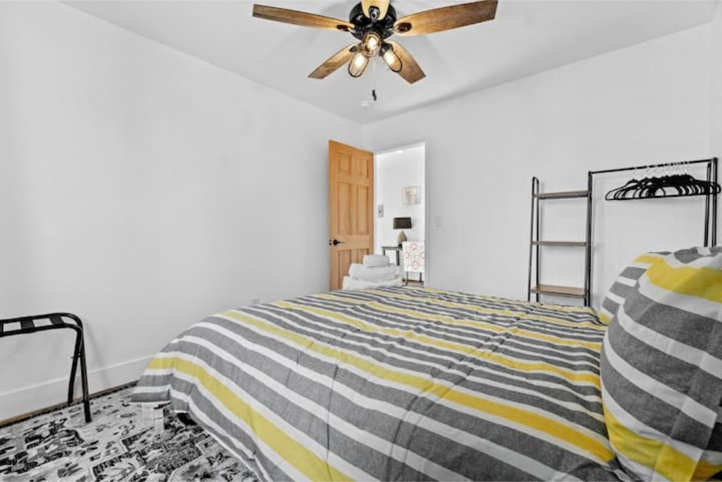 A bedroom with a striped bedspread, a ceiling fan, a wooden door, a metal rack, and a black and white patterned rug.