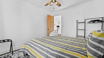 A bedroom with a striped bedspread, a ceiling fan, a wooden door, a metal rack, and a black and white patterned rug.