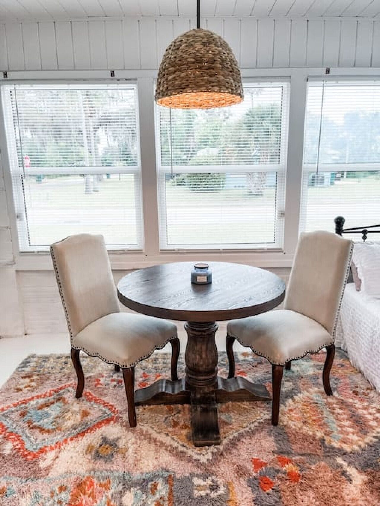 A round wooden table with two upholstered chairs, a woven pendant light, and a colorful rug.