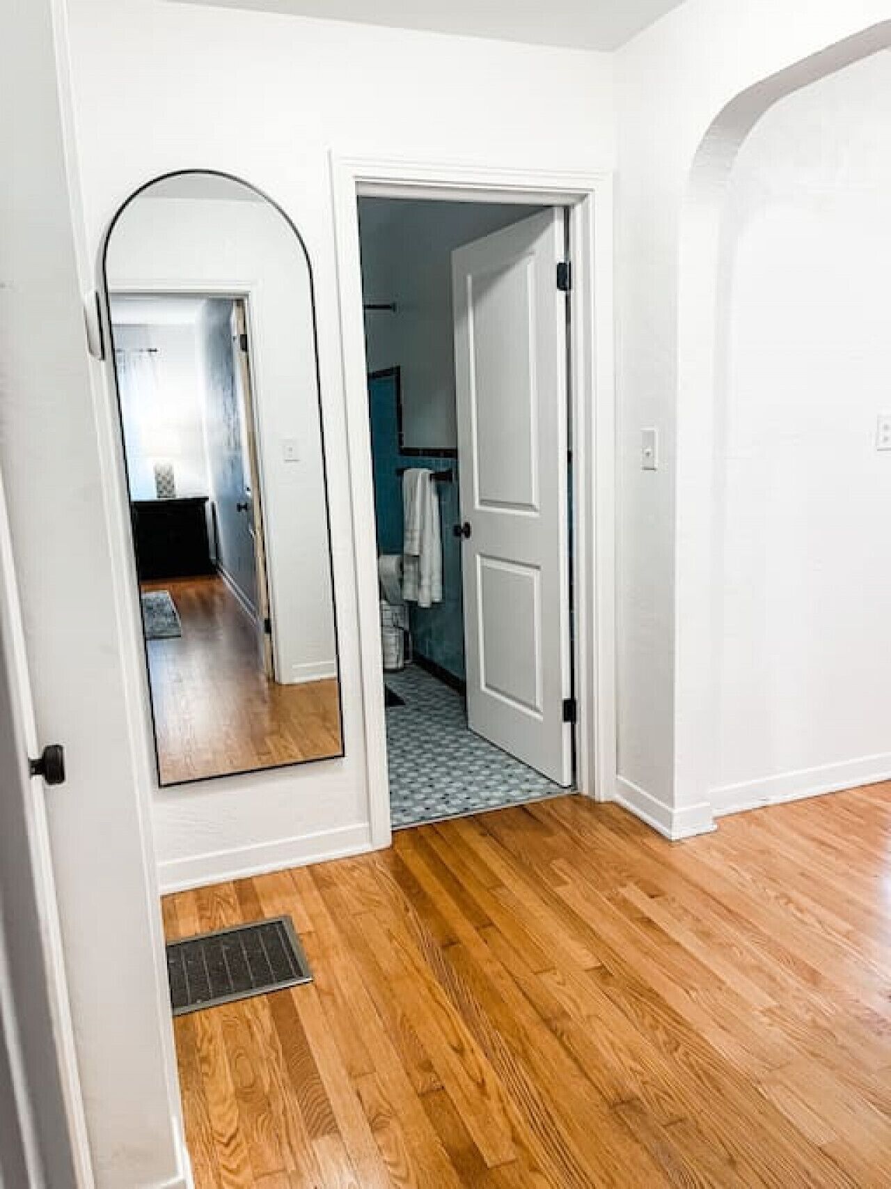 A hallway with wooden flooring, white walls, and a mirror on the left.