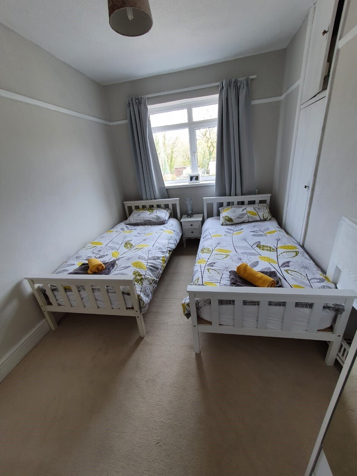 A small, well-lit room with two single beds, a window with curtains, and a white wardrobe.