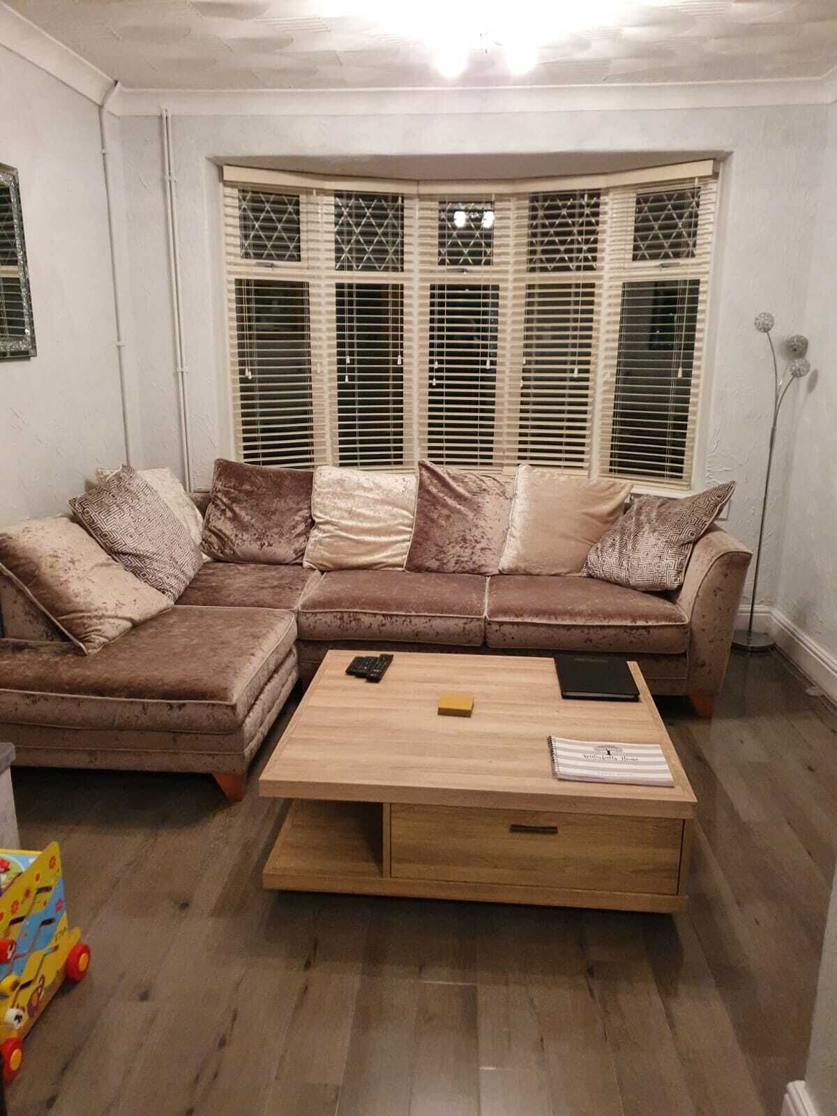 Un salon chaleureux avec un canapé d’angle marron, une table basse en bois et une baie vitrée équipée de stores.