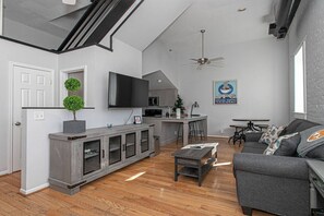 A modern living room with a grey sofa, a coffee table, and a TV. There's a kitchen area with a microwave and a ceiling fan.