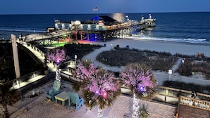 A pier extending into the ocean with a building at the end, a sandy beach area with pathways, and a small boat docked nearby. - Paramount Plus/Oceanfront/New (Myrtle Beach)