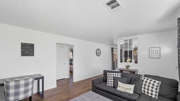 A modern living room with a grey sofa, a plaid armchair, and a grey ottoman. There is a black and white checkered pillow on the sofa. A black sign on the wall reads "WELCOME TO TULARE ENJOY OR STAY". - The Cozy Rustic Sequoia Wi-Fi & 2 Garage (Tulare)