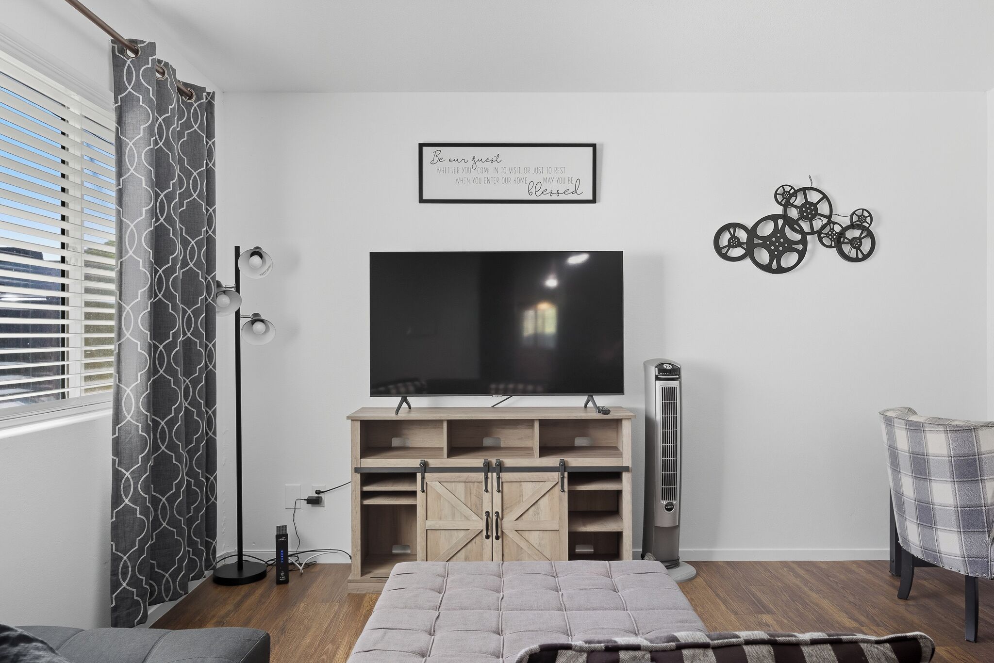 Ein Wohnzimmer mit einem großen Flachbildfernseher, einer Holz-Unterhaltungseinrichtung, einer Stehlampe, einem Standheizkörper und einem karierten Sessel.