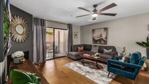 A modern living room with a sofa, coffee table, and a blue armchair. There is a ceiling fan, a large mirror, and a painting on the wall.