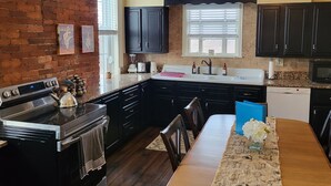 A kitchen with a brick wall, dark cabinets, a wooden dining table, and a ceiling fan.