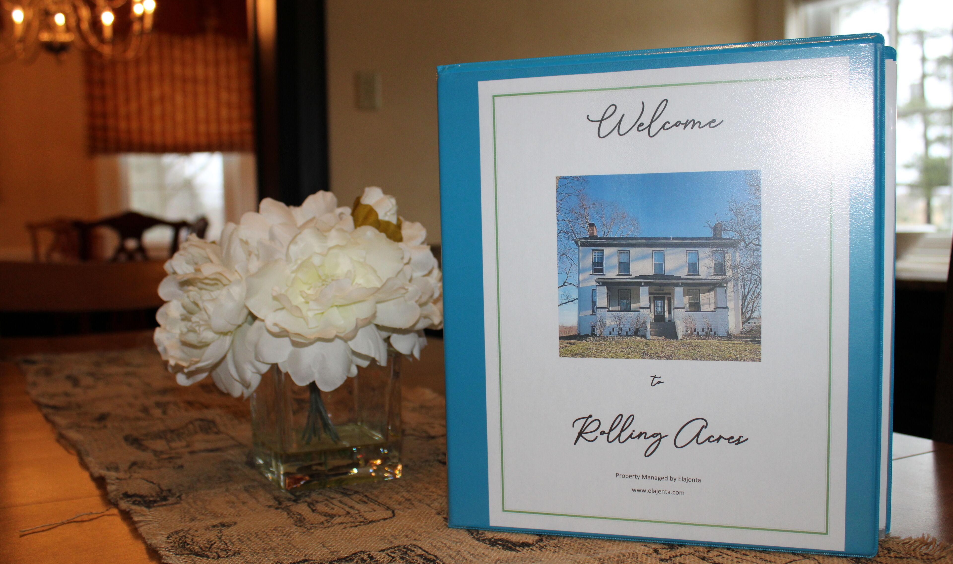 A framed welcome sign with a house photo and text, a vase of white flowers, and a patterned tablecloth.