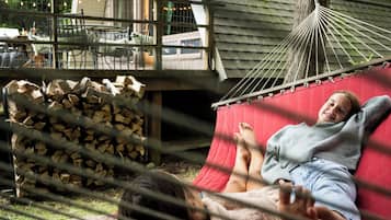 Un hamac dans lequel est allongé une personne, une pile de bois de chauffage et, au fond, un bâtiment doté d’un toit en pente.