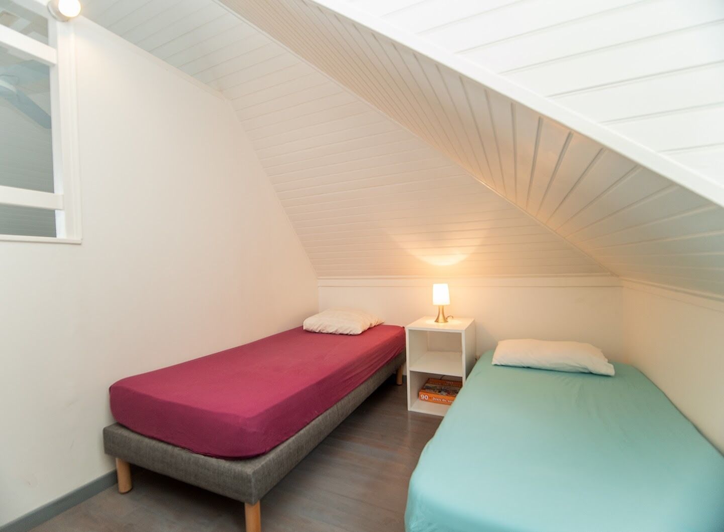 A small, attic-style room with two single beds, each with a different colored mattress. There is a nightstand with a lamp between the beds.