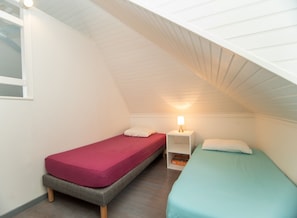 Une petite chambre aménagée sous les combles, avec deux lits simples, chacun doté d’un matelas de couleur différente. Une table de chevet avec une lampe se trouve entre les deux lits.