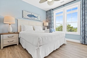A bedroom with a large bed, two bedside tables with lamps, a ceiling fan, a window with curtains, and a wall-mounted artwork of a fish.