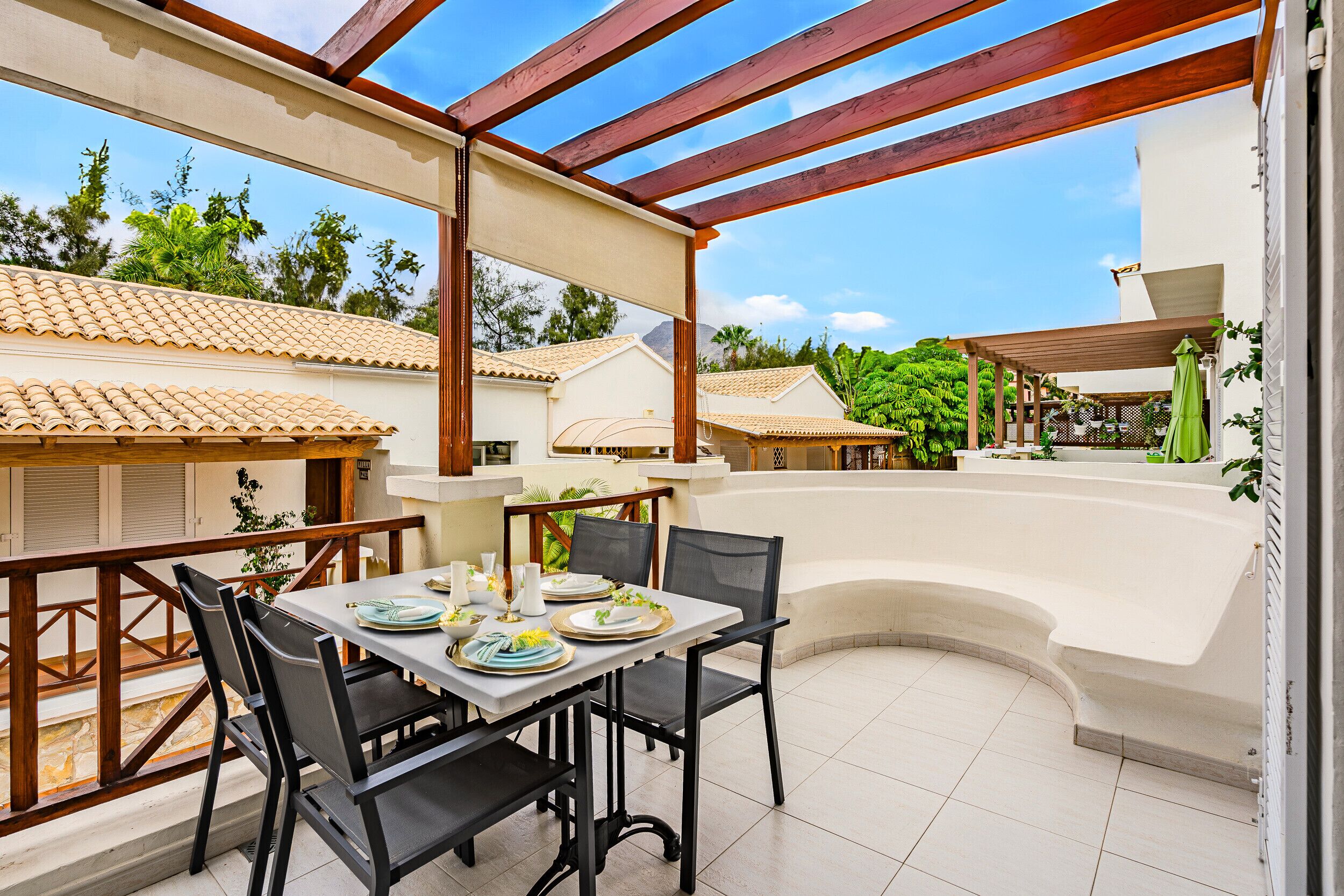 A covered outdoor dining area with a table set for two, surrounded by chairs, and a curved bench.