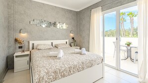 Un dormitorio moderno con una cama grande, una mesita de noche, una lámpara de pared y un ventanal corredero que da a un espacio exterior con una mesa y sillas.