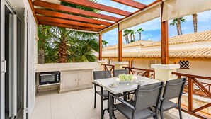 Un patio exterior con una pérgola de madera, una mesa con sillas, un microondas y una pequeña zona de cocina.