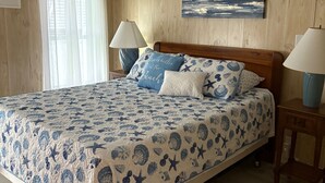A bedroom with a wooden paneled wall, a bed with a blue and white beach-themed bedspread, two bedside lamps, a ceiling fan, and a painting of a beach scene. - Sunshine and Relax! (Zephyr Hill)