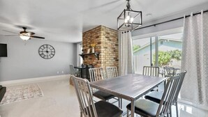 A dining area with a brick fireplace, a ceiling fan, and a large window with curtains.